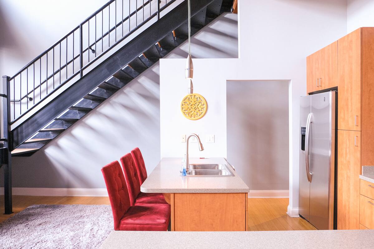 Open-concept urban loft kitchen with black metal staircase, light wood cabinets, stainless steel refrigerator, center island sink and two red velvet counter chairs