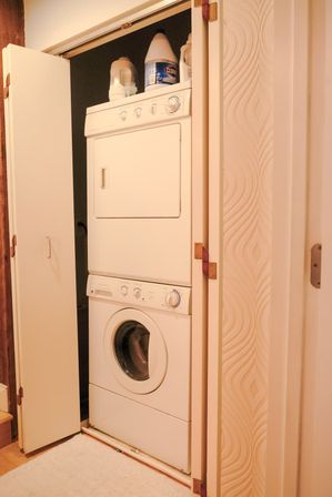 Compact white stacked washer-dryer tucked into a narrow hallway laundry closet with bi-fold doors open and detergent jugs on top.
