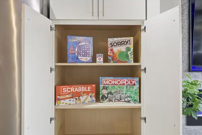 Open white cabinet in a family room displaying board games for game night — Scrabble, Monopoly, Sorry!, Connect 4 and a deck of cards.