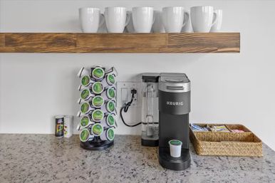 Kitchen countertop coffee station with a black single‑serve pod brewer, rotating coffee pod rack, wicker caddy of sweeteners and white ceramic mugs on a wooden floating shelf over a speckled granite counter.