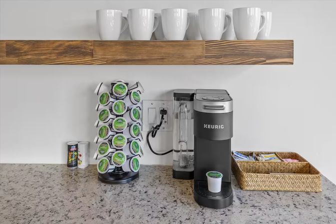 Kitchen countertop coffee station with a black single‑serve pod brewer, rotating coffee pod rack, wicker caddy of sweeteners and white ceramic mugs on a wooden floating shelf over a speckled granite counter.