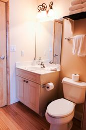Cozy small bathroom with white vanity and sink, toilet with spare roll, wall mirror, folded towels on a shelf, and warm wood flooring.