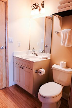 Cozy small bathroom with white vanity and sink, toilet with spare roll, wall mirror, folded towels on a shelf, and warm wood flooring.
