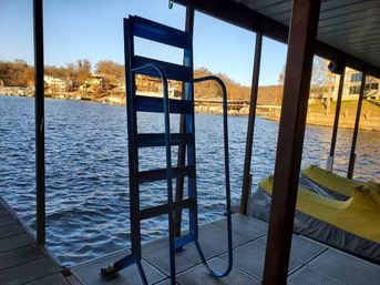 Blue metal ladder on a covered lake dock beside yellow-covered jet skis, overlooking rippling lake water and lakeside homes.