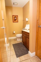 Cozy small bathroom with tan walls, beige tile floor, dark gray bath mat, white toilet, wooden vanity topped with towels, and framed state-shaped wall art.