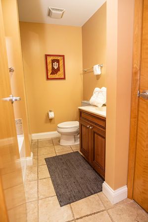 Cozy small bathroom with tan walls, beige tile floor, dark gray bath mat, white toilet, wooden vanity topped with towels, and framed state-shaped wall art.