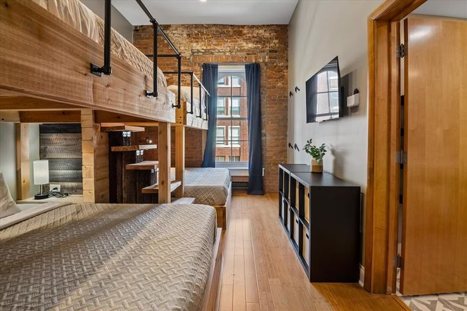 Cozy urban loft-style bedroom with wooden bunk beds and lower double beds, exposed brick wall, hardwood floors, tall window with navy curtains, industrial metal railings, wall-mounted TV, black cubby storage and small potted plant.