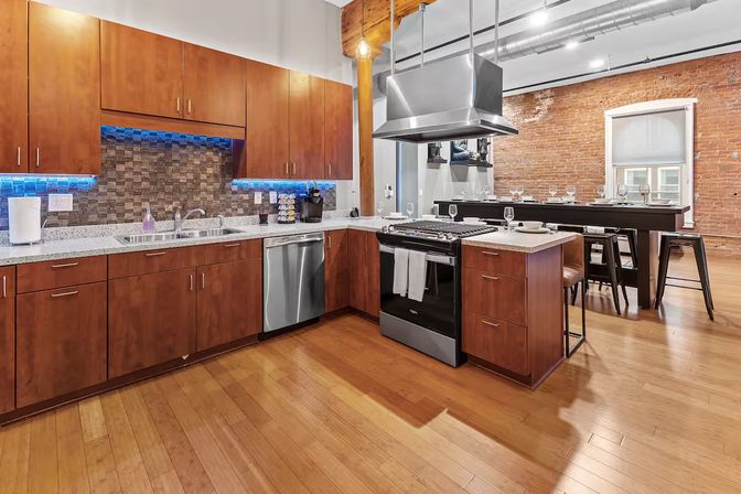 Modern urban loft kitchen with warm wood cabinets, stainless steel range and dishwasher, stone tile backsplash with blue LED lighting, center island and dining bar against exposed brick and hardwood floors.