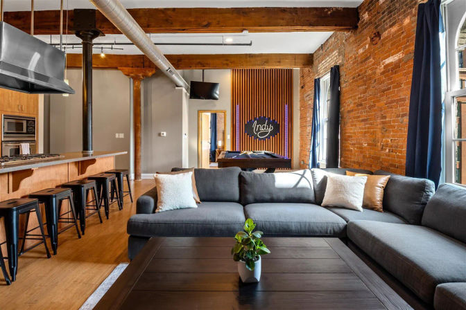 Bright urban loft living room with exposed brick walls and wooden beams, large gray sectional and dark wood coffee table with small plant, open kitchen island with black metal stools, and a pool table under a decorative "Indy" wall sign.