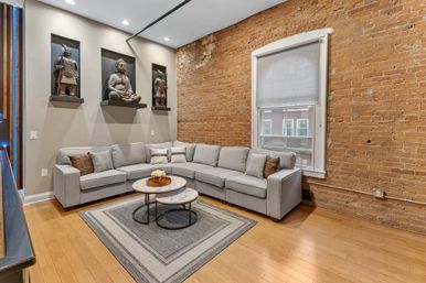 Cozy urban loft living room with exposed brick wall, large gray L-shaped sectional sofa, nested round coffee tables on a geometric rug, recessed Buddha and warrior statues in wall niches, hardwood floors and a window with a roller shade.