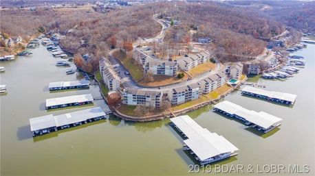 Aerial view of lakefront condominiums on a curving wooded peninsula with multiple covered marinas and boat slips moored on calm water, late‑fall trees and winding roads