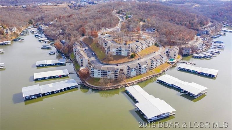 Aerial view of lakefront condominiums on a curving wooded peninsula with multiple covered marinas and boat slips moored on calm water, late‑fall trees and winding roads