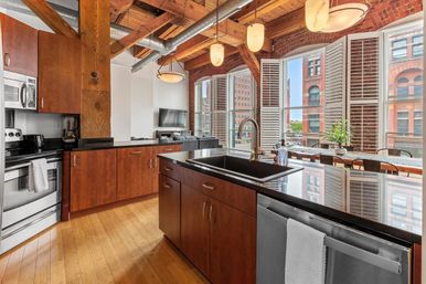 Urban loft kitchen with exposed wooden beams and brick walls, arched bay windows offering city views, black granite island with sink, stainless appliances and warm wood cabinetry.