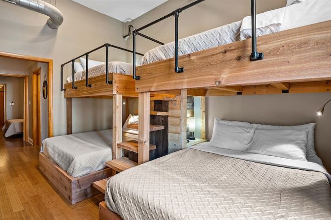 Cozy cabin-style bunk room with built-in wooden loft beds (two lower full beds and two upper bunks), black metal railings, neutral quilted bedding, bedside lamp and warm hardwood floors — family-friendly vacation rental vibe.