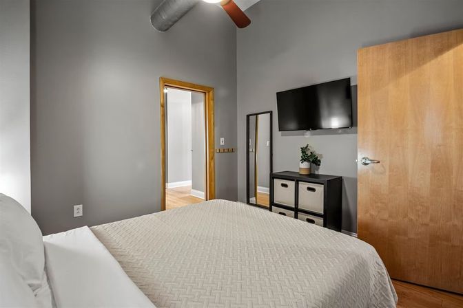 Cozy modern urban loft bedroom with gray walls, exposed ductwork, bed topped with a textured beige quilt, wall-mounted TV, full-length mirror, black cube dresser with fabric bins, and wooden door.