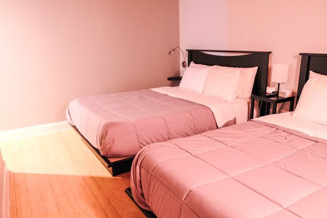 Cozy minimalist guest room with two double beds in soft pink comforters, black headboards, bedside table with lamp, and warm hardwood floor