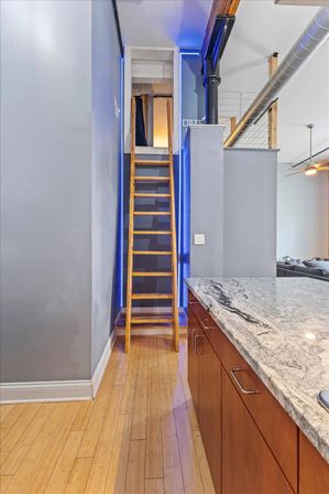 Modern urban loft interior with a wooden ladder to a mezzanine lit by blue LED strips, granite kitchen island, warm bamboo floors and exposed ductwork.