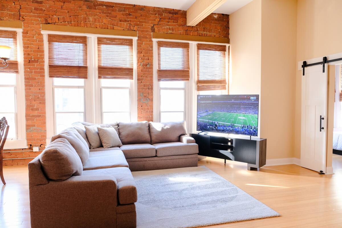 Cozy sunlit urban loft living room with exposed brick wall, four tall windows with bamboo blinds, gray L-shaped sectional sofa on a light area rug, hardwood floors, flat-screen TV showing a football game on a modern media console, and a sliding barn door to the bedroom.