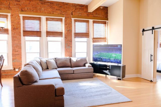 Cozy sunlit urban loft living room with exposed brick wall, four tall windows with bamboo blinds, gray L-shaped sectional sofa on a light area rug, hardwood floors, flat-screen TV showing a football game on a modern media console, and a sliding barn door to the bedroom.