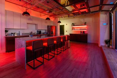 Modern urban loft kitchen and bar with a long granite island, six red-upholstered bar stools, hanging pendant lights, stainless refrigerator, hardwood floors and moody red LED accent lighting under the island and on wall shelves.