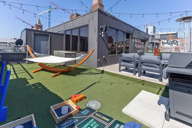 Urban rooftop patio with a wooden hammock, artificial turf, gray lounge seating, outdoor grill, string lights and construction cranes in the skyline.