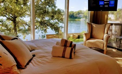 Sunlit lakefront bedroom with made bed and rolled towels, cozy armchair and floor-to-ceiling windows offering a panoramic view of a tree-lined lake and dock