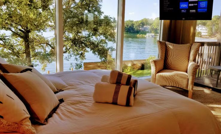 Sunlit lakefront bedroom with made bed and rolled towels, cozy armchair and floor-to-ceiling windows offering a panoramic view of a tree-lined lake and dock