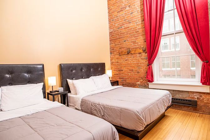 Cozy urban loft-style hotel room with two beds, tufted dark headboards, exposed red brick wall, hardwood floors, bedside lamps and a large window with red curtains overlooking city buildings.