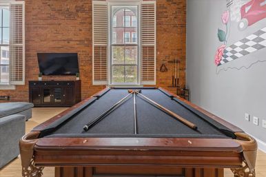 Black-felt pool table with crossed cues in an urban loft game room — exposed brick wall, arched shuttered window, TV cabinet, wall mural and hardwood floors.