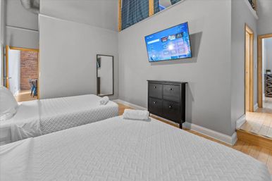 Modern urban loft bedroom with two white‑lined beds, wall‑mounted TV, black dresser, full‑length mirror, hardwood floors and exposed brick visible through doorway.