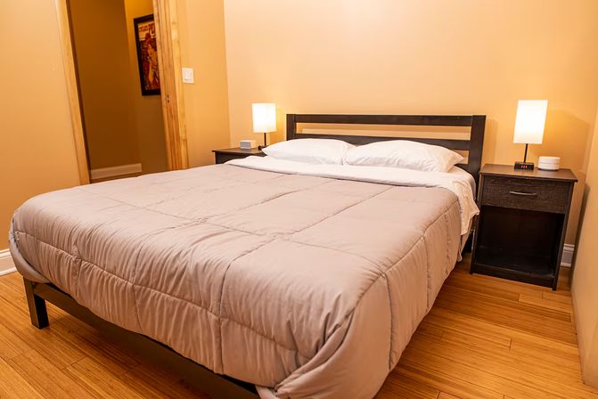 Cozy bedroom with neatly made queen bed, beige comforter, dark wood headboard and nightstands, warm bedside lamps and hardwood floor.