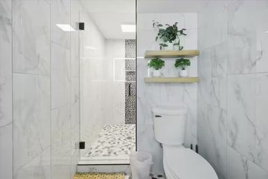 Bright modern marble bathroom with a frameless glass shower and pebble-tile floor, white toilet and wooden floating shelves holding green potted plants.