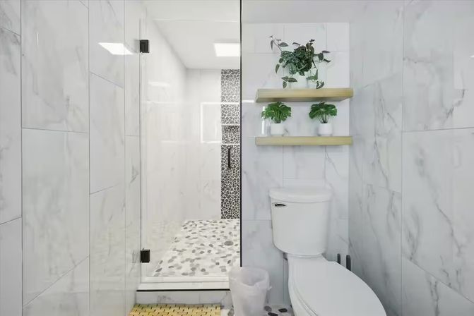 Bright modern marble bathroom with a frameless glass shower and pebble-tile floor, white toilet and wooden floating shelves holding green potted plants.