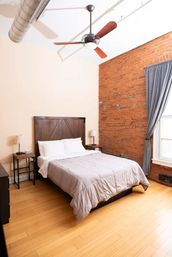 Cozy urban loft bedroom with exposed red brick wall, wooden headboard and queen bed dressed in a gray comforter, hardwood floors, bedside tables with lamps, ceiling fan, and a tall window with gray curtains.