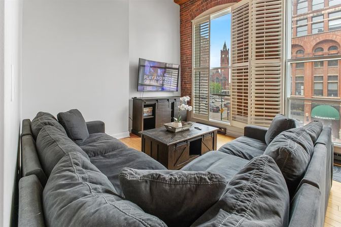 Bright urban loft living room with exposed brick wall and large windows with plantation shutters overlooking a historic brick clock tower, furnished with a plush gray sectional, wooden coffee table, TV console and hardwood floors.