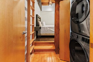 Cozy studio loft hallway with warm hardwood floors, stacked washer and dryer in a laundry closet, wooden ladder and steps leading up to a minimalist bed beneath exposed ceiling beams.