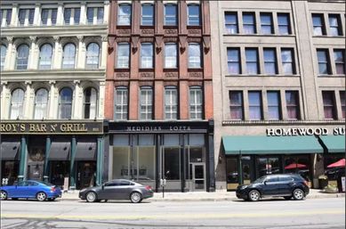 Downtown streetscape of three adjacent historic commercial buildings – cream, red-brick and tan facades with arched windows, street-level storefronts and parked cars along the curb.