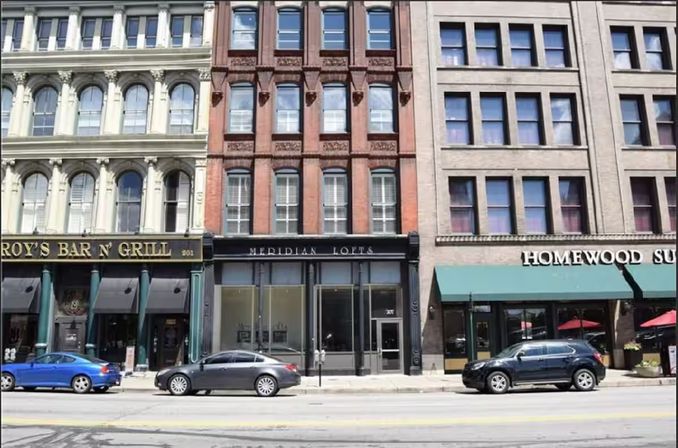 Downtown streetscape of three adjacent historic commercial buildings – cream, red-brick and tan facades with arched windows, street-level storefronts and parked cars along the curb.