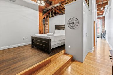 Sunny industrial loft bedroom on a raised platform with a dark wooden bed, exposed timber beams and brick, hardwood floors, metal ductwork, wall clock and a long hallway with shuttered windows.