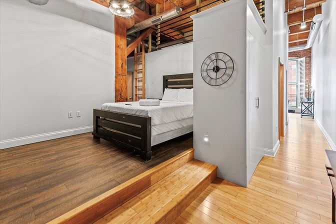 Sunny industrial loft bedroom on a raised platform with a dark wooden bed, exposed timber beams and brick, hardwood floors, metal ductwork, wall clock and a long hallway with shuttered windows.