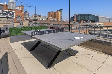 Sunlit rooftop ping-pong table on a modern urban terrace with paddles, planters, string lights and downtown city skyline in the background.
