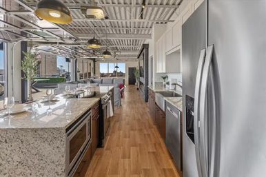 Open-plan urban loft kitchen with granite island, stainless steel appliances, pendant lights, wood floors and folding glass doors opening to a rooftop terrace and city skyline.