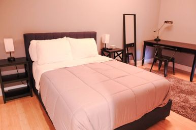 Cozy modern bedroom with upholstered headboard, blush comforter and white pillows, black nightstands and lamps, full-length mirror, desk and chair on hardwood floor.