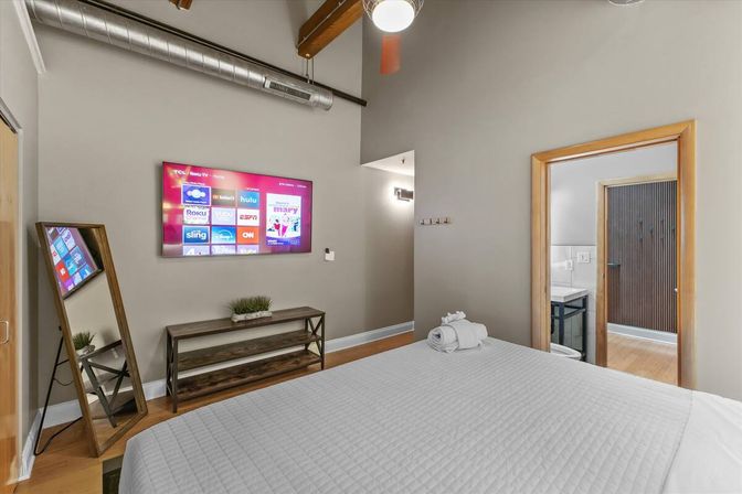 Cozy modern loft bedroom with a white-quilted bed, wall-mounted smart TV showing streaming apps, leaning full-length mirror, wooden console, exposed metal ductwork and doorway to an en-suite bathroom.