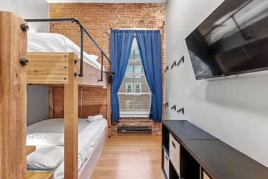 Cozy industrial loft bedroom with wooden bunk beds, exposed brick wall, tall window framed by blue curtains overlooking city buildings, wall-mounted TV, black storage shelves and hardwood floor.