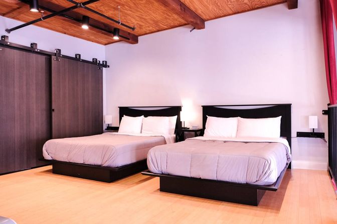Bright urban loft-style guest room with exposed wooden ceiling beams, sliding barn-door closet, hardwood floor, and two queen beds with gray duvets and bedside lamps.