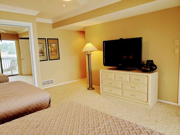 Cozy carpeted bedroom with two quilted beds in the foreground, a flat-screen TV on a cream dresser, a tall floor lamp and framed art on warm beige walls, plus a sliding door to a balcony with a water view.