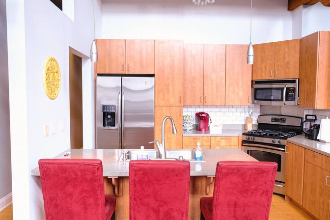 Bright modern kitchen with a central island, three red upholstered bar stools, stainless steel refrigerator and range, wood cabinets, subway-tile backsplash, pendant lights and a red coffee maker.