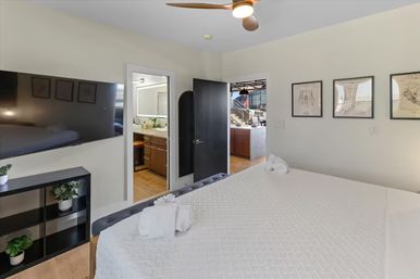 Bright modern urban loft bedroom with white quilted bed and folded towels, wall-mounted flat-screen TV, black shelving with potted plants, ceiling fan, framed art, and open doorways to an en-suite bathroom and open kitchen