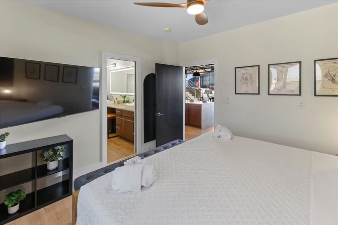 Bright modern urban loft bedroom with white quilted bed and folded towels, wall-mounted flat-screen TV, black shelving with potted plants, ceiling fan, framed art, and open doorways to an en-suite bathroom and open kitchen
